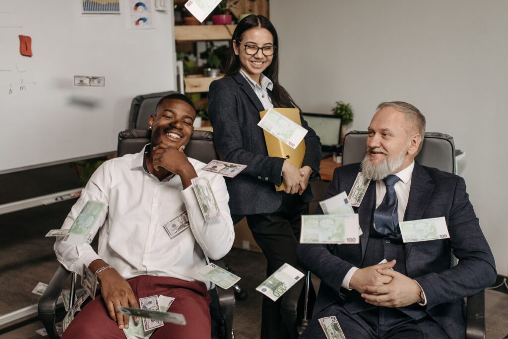 Three colleagues laughing in an office as banknotes fall around them