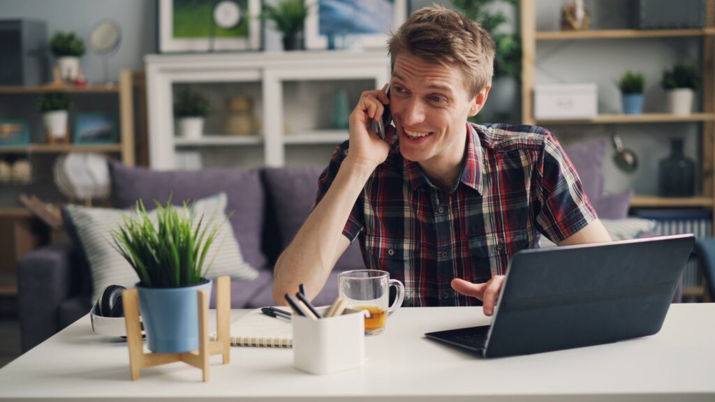 Man on a phone call while working at a laptop in a bright home office